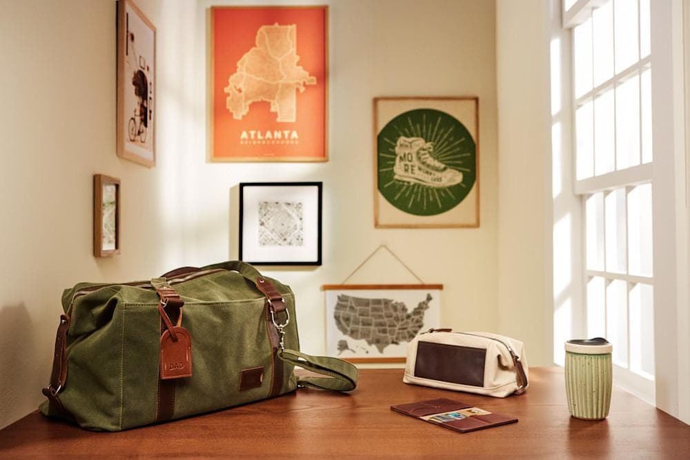 A duffle bag, dopp kit and ceramic mug sit atop a wood table in front of a wall of art prints.
